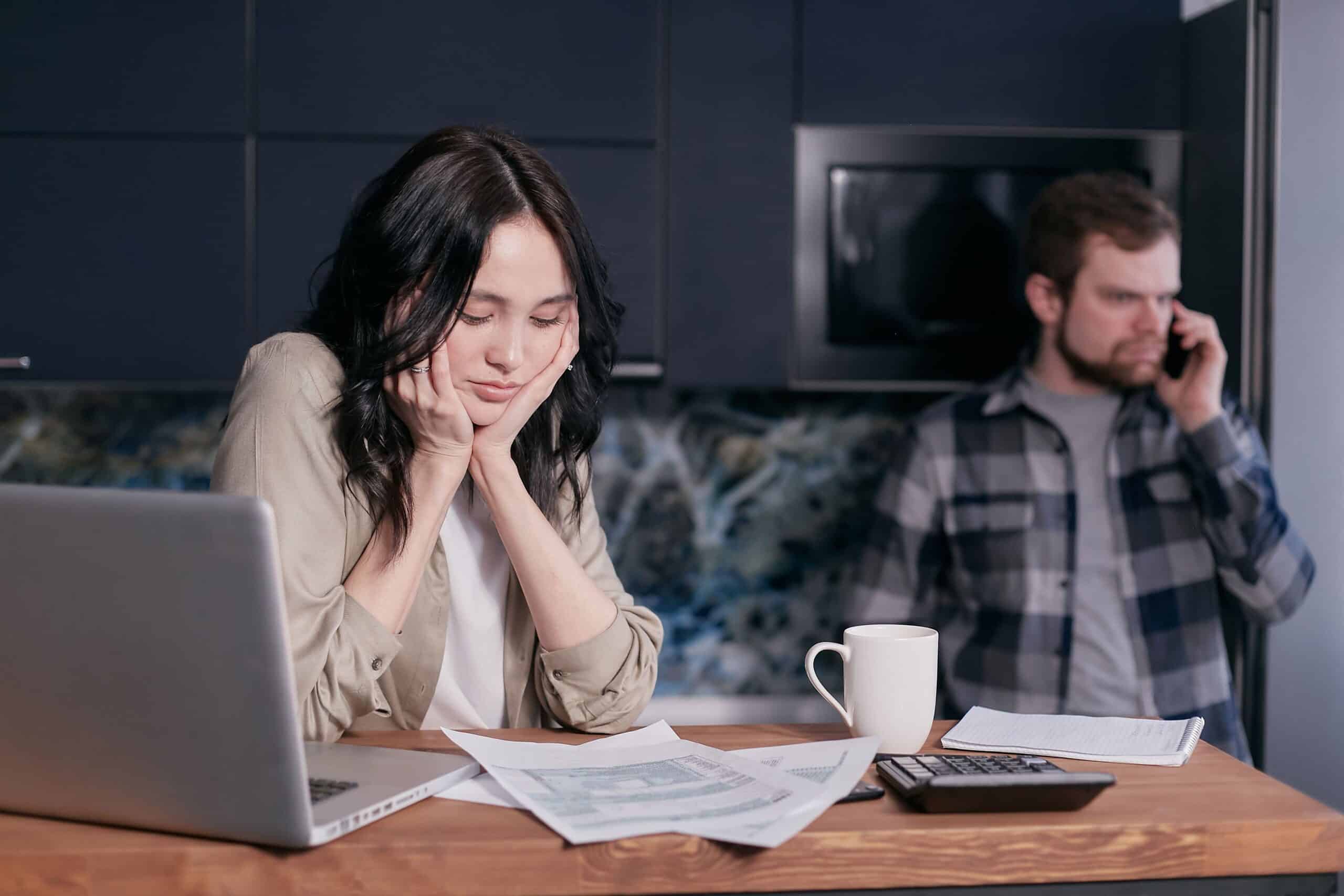 a woman sits at a kitchen table with an open laptop and paperwork in front of her. a man in the background looks concerned and is on the phone.